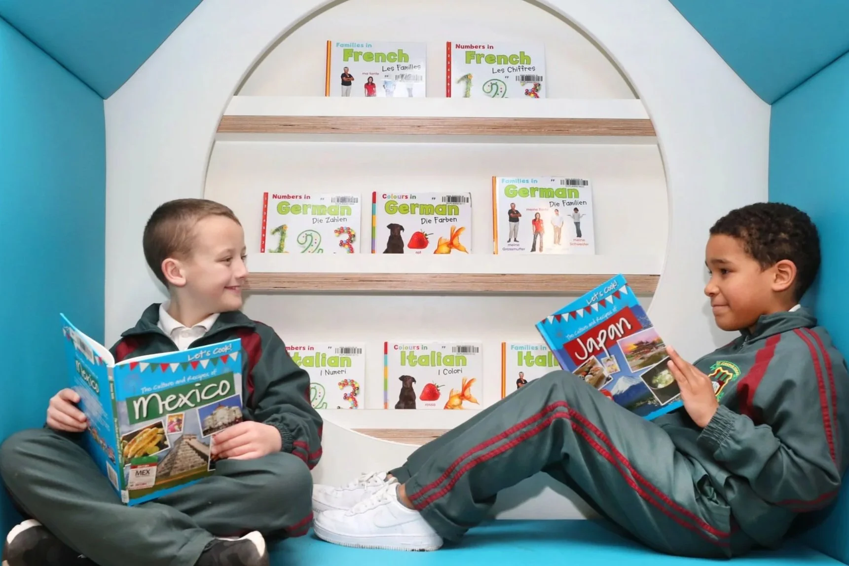 Two boys in school uniforms sitting inside a small, enclosed bookshelf reading travel books about Mexico and Japan.