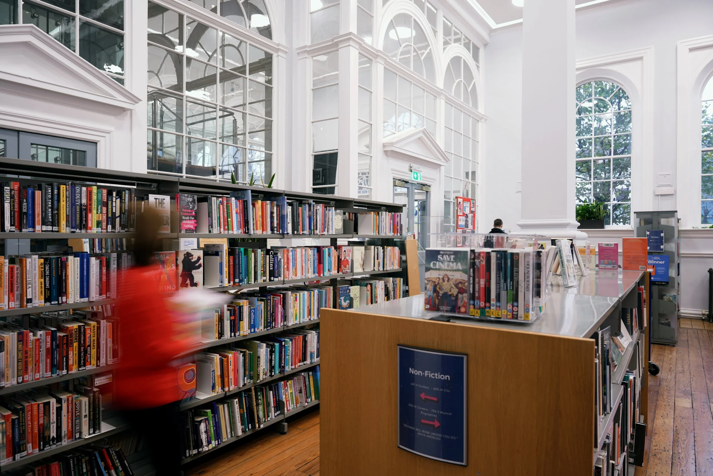 Interior of a bright, spacious library with tall white walls and large arched windows, filled with bookshelves and a table with books, with a blurred person walking past.