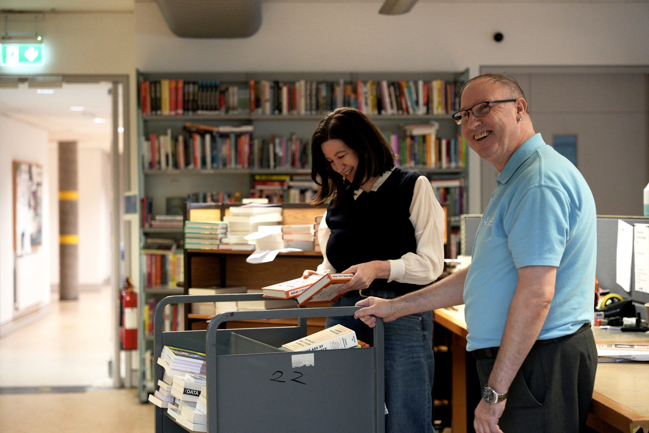 Two people, a woman with dark hair and a man with glasses, smiling and looking at books in a library or bookstore. They are standing by a cart filled with books, with bookshelves in the background.