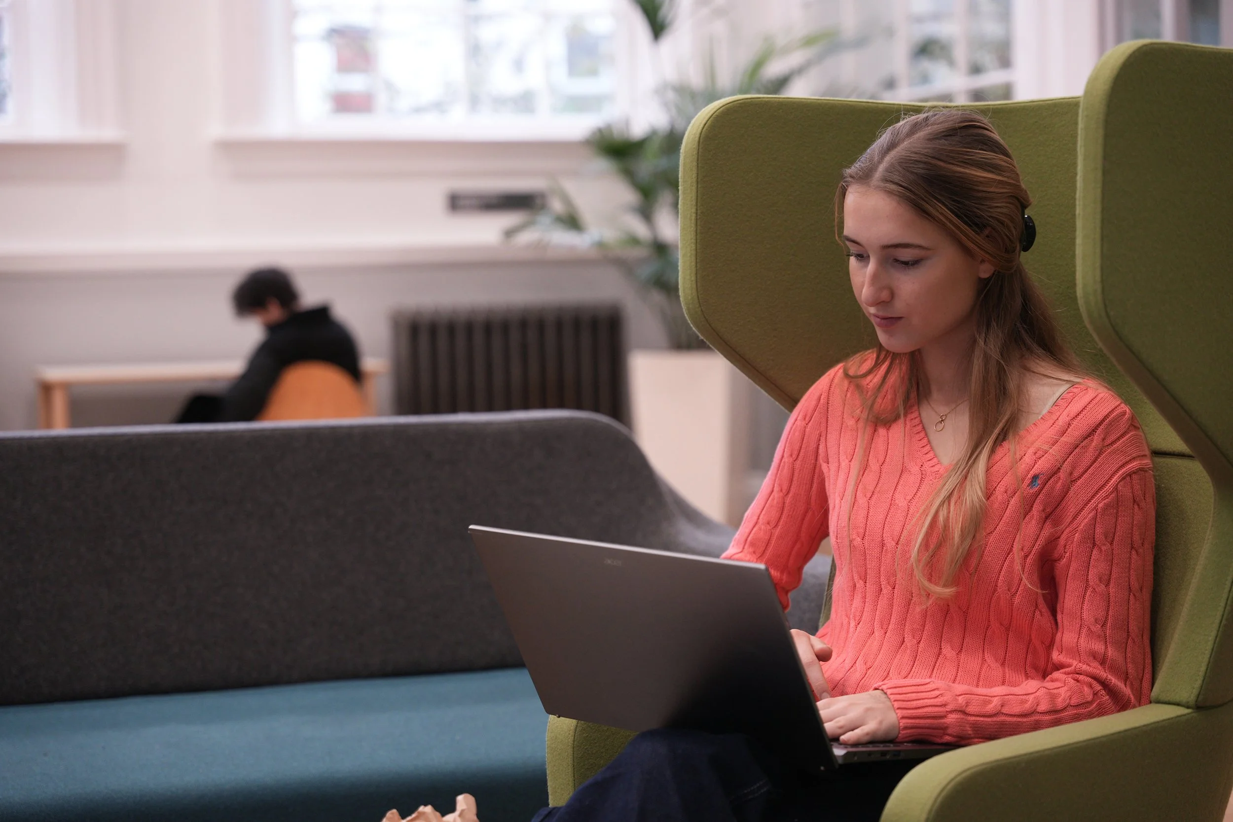 A young woman with long blonde hair, wearing a coral sweater, sitting on a modern green chair with a laptop on her lap, in a bright indoor space with large windows and a potted plant in the background.