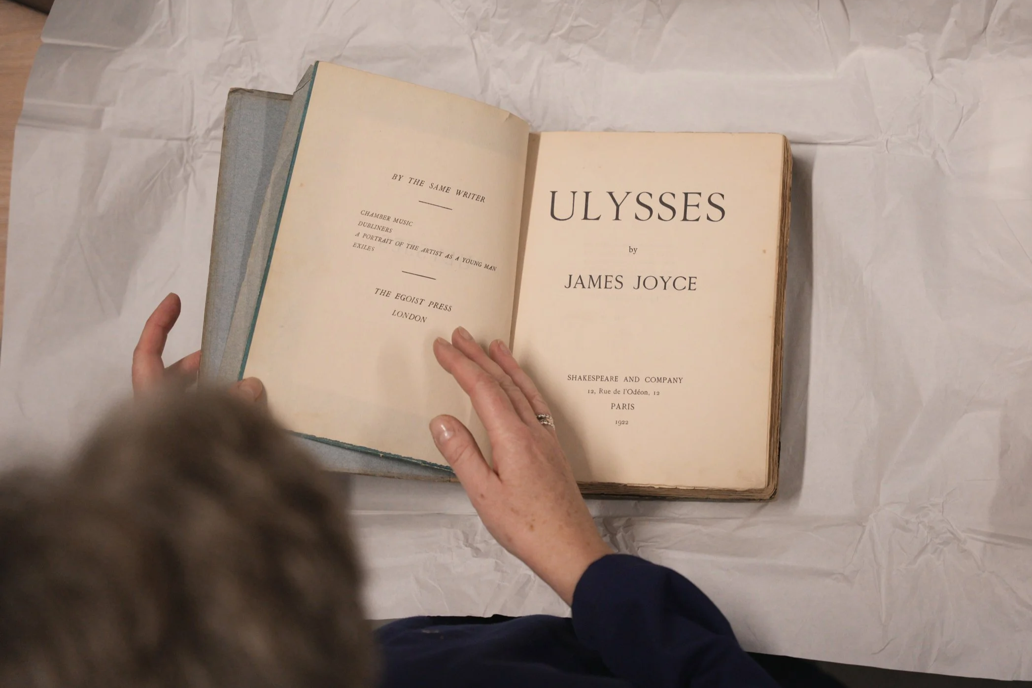 An open vintage book titled "Ulysses" by James Joyce displayed on crumpled paper, with a person's hand resting on the right page, seen from above.
