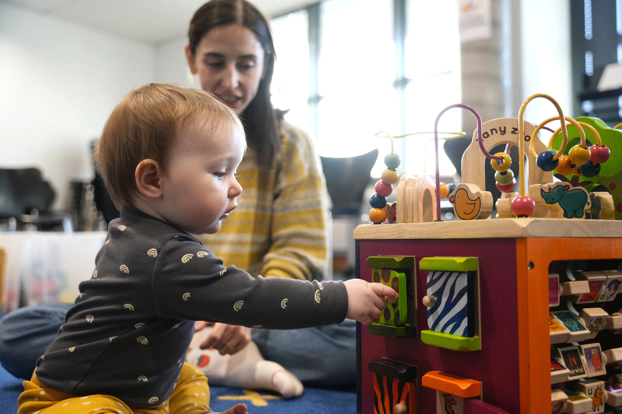 A young child in a dark gray shirt with rainbow patterns and yellow pants playing with a colorful wooden activity cube, with a woman in a yellow striped shirt observing in the background, in a room with bright natural light.