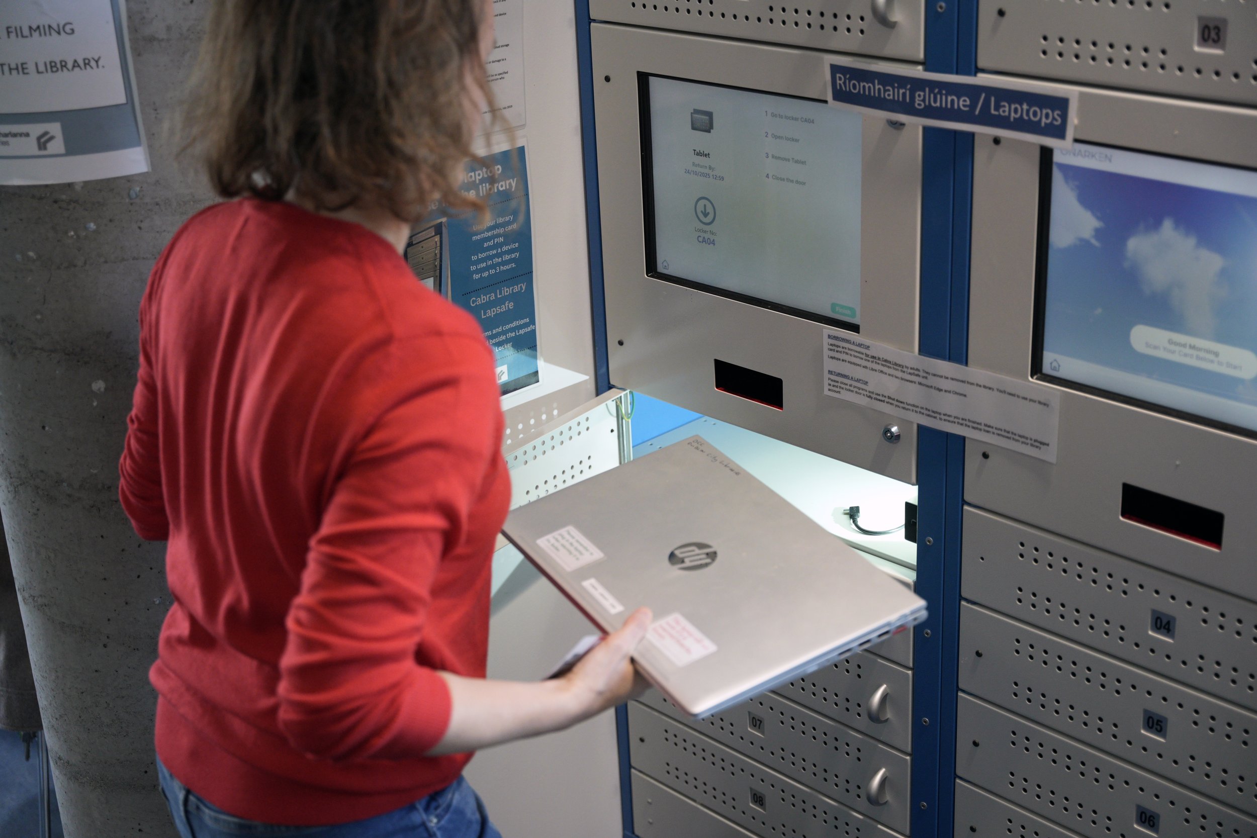A person in a red shirt using a locker to pick up a laptop in a library locker station.