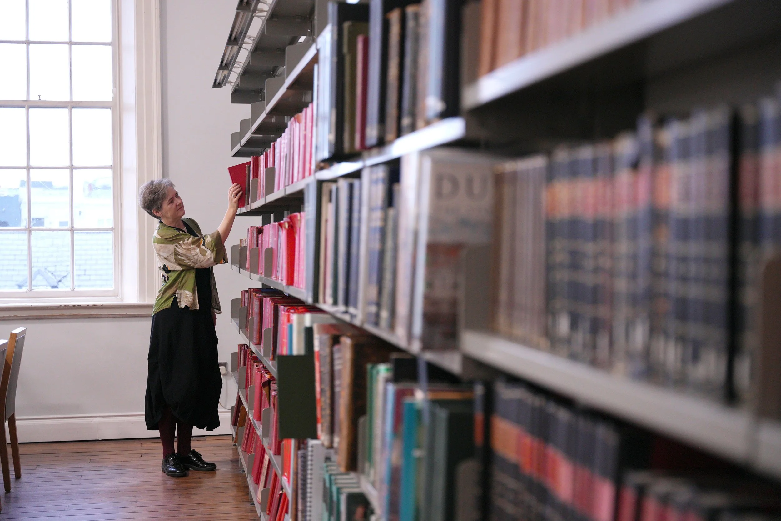 An older woman browsing books on a tall bookshelf in a library, with a large window in the background.