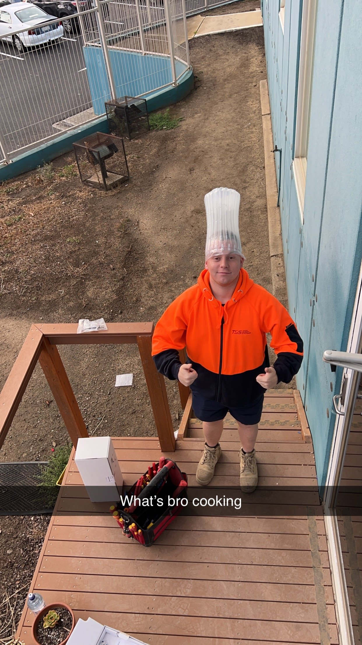 A young man in an orange jacket, black shorts, beige boots, and a plastic chef's hat is standing on a small wooden porch steps, smiling and pointing with both fists. There are various objects on the porch including a box, a container of tools, a water bottle, and some papers. Behind him, there is a blue building with a window, an outdoor dirt area, and a parking lot with cars.