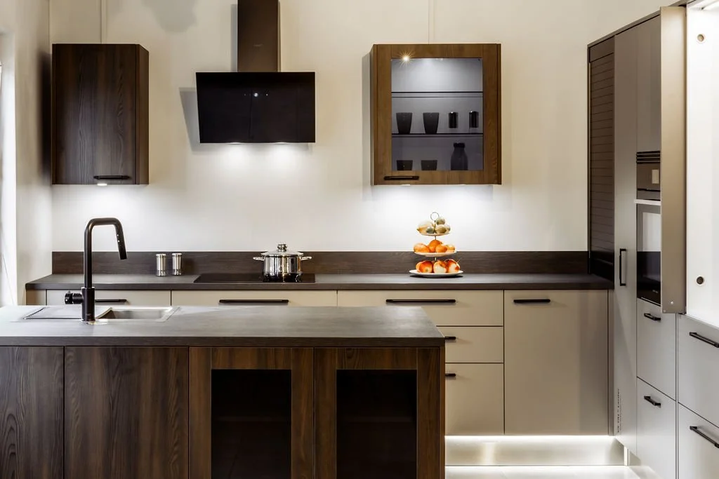 Moderni keittiö, jossa tummat ja vaaleat kaapistot, pesuallas ja nestelevyt työtasolla, koriste-esineitä ja astioita näytillä.