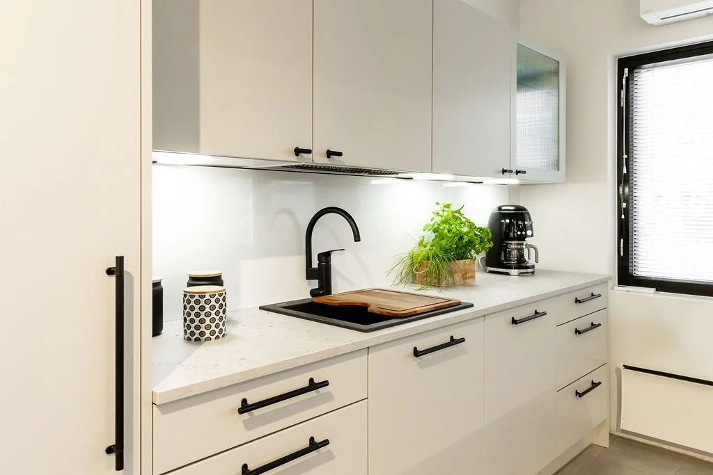 Valkoinen keittiö, jossa musta hana, pottipuutarha, kahvinkeitin ja keittiövälineitä, ikkunassa mustat kaihtimet