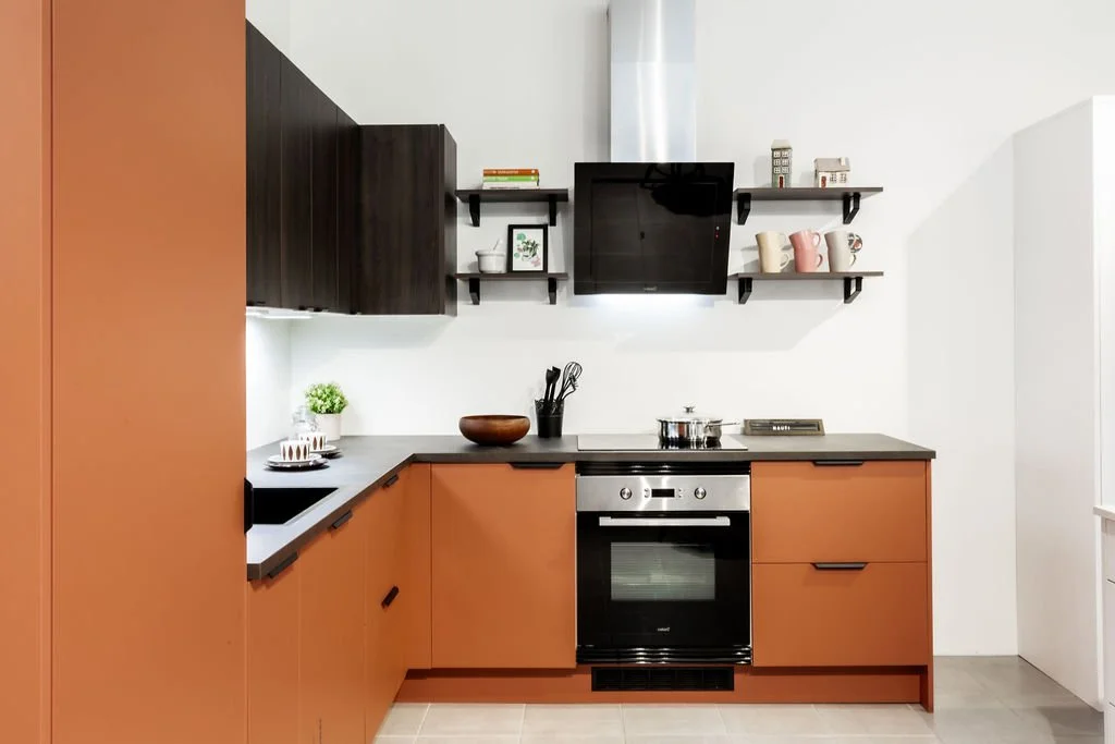 Kodin keittiö, jossa vaaleanruskeat alakaapit, musta työtaso, liesi ja uuni, mustat seinähyllyt, pieni vihreä kasvi, keittiövälineitä ja astioita.