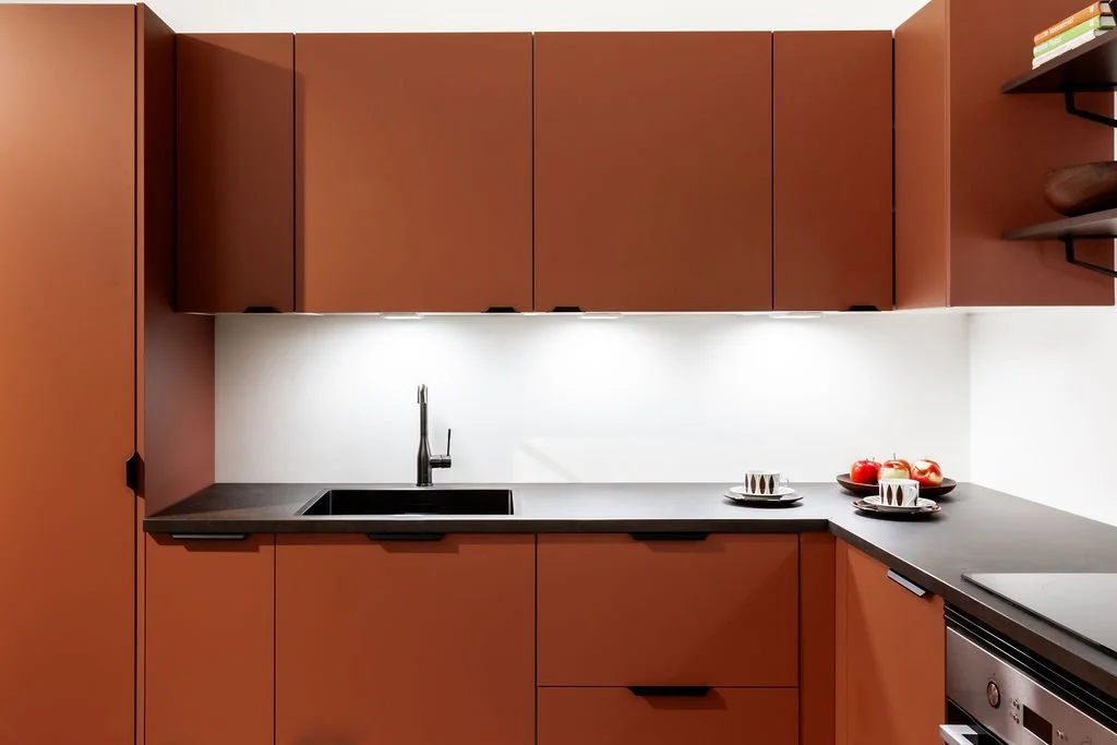 Keittiö, jossa ruskea kaappi ja musta työtaso, liesi oikealla ja lavuaari keskellä, valkoiseksi maalattu seinä taustalla, keittiössä on kaksi lautasen ja kolme omenaa kulhoissa.