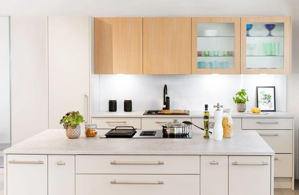 Moderni keittiö, jossa valkoinen saareke, puinen yläkaapit ja valkoiset alakaapit, pieniä kasveja, keittiövälineitä ja koriste-esineitä.