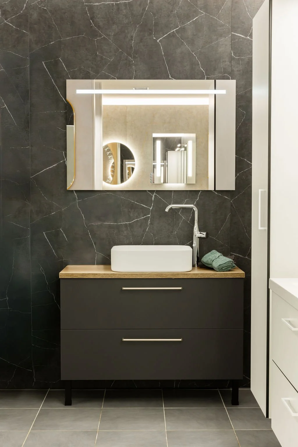 Kohokuvioinen tumma seinä ja moderni kylpyhuoneen allaskaappi, yläpuolella peili ja valoisat valaistuslaitteet.