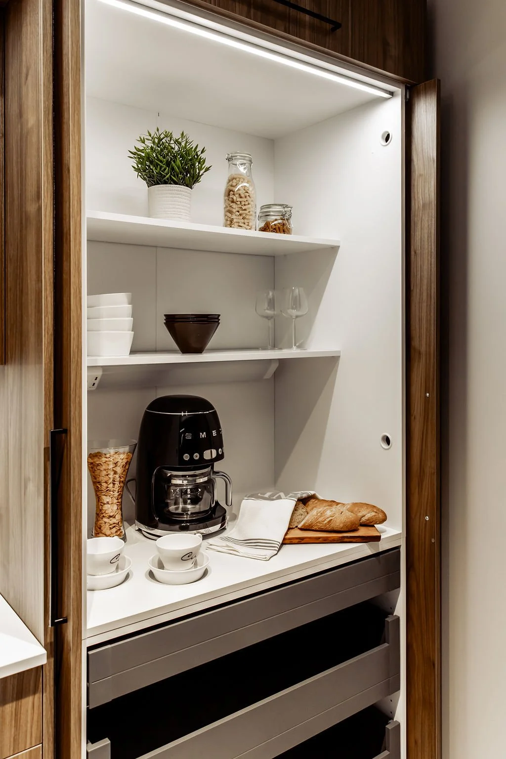 Keittiön kaapisto, jossa on kahvi- ja leivontatarvikkeita, kuten kahvinkeitin, leipiä, lasiastiasto, kastikkeita ja kasvi.