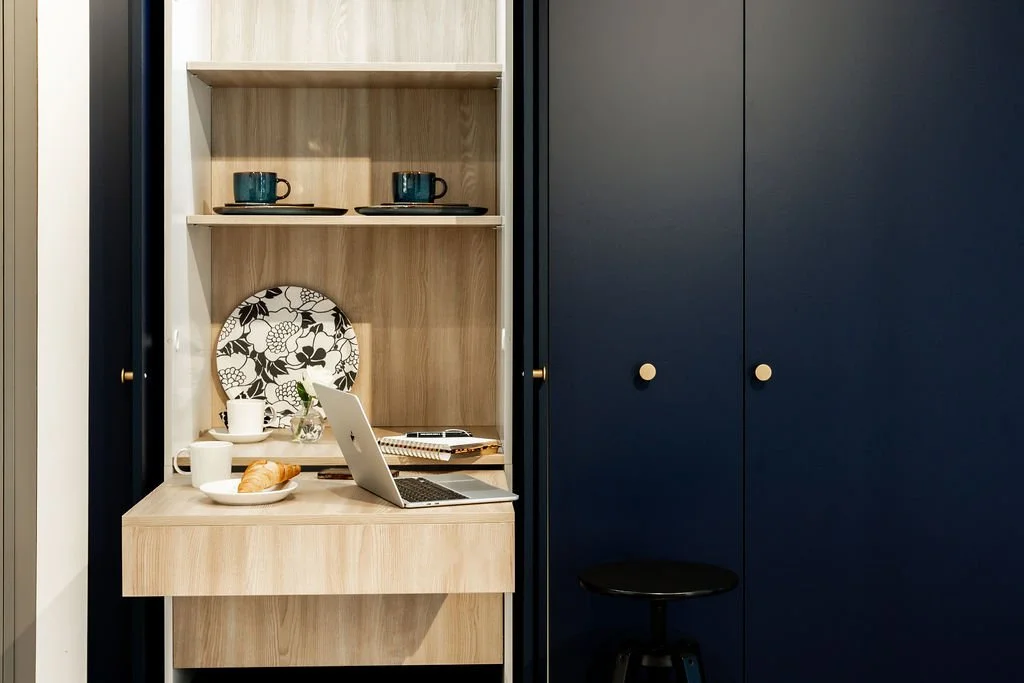 Kuvan esityksessä on kodikas keittiön tai ruokailutilan nurkkaus, jossa on avoin puinen hylly ja pieni työpöytä, jossa on kannettava tietokone, kahvikuppi, leivonnainen ja kirjoja.