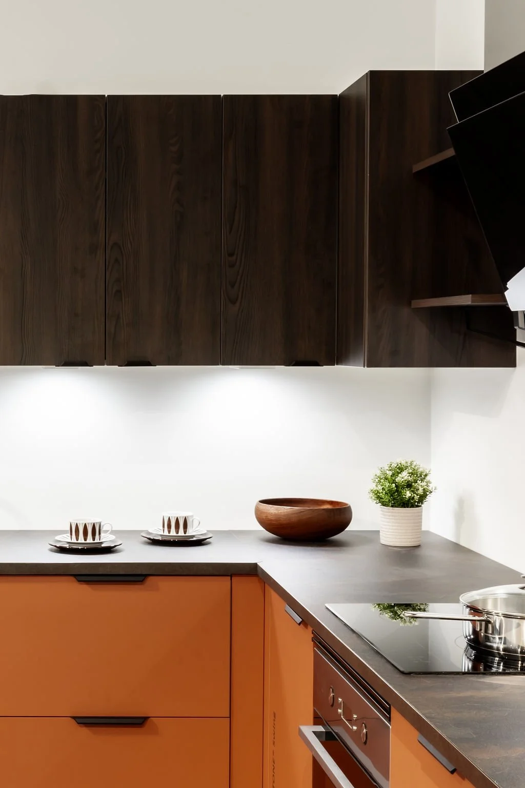 Keittiö, jossa ruskeat ylähyskompien, oranssit alahyllyt ja tummanharmaa työpöytä, lavara tanko, maljakko ja kasvi.