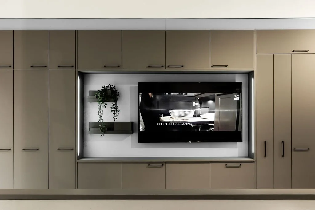 Moderni televisio seinällä, ympäröi valkoinen valaistu kolo, hyllyt ja pienet suojaruukut, beige kaapit ja kaapistot ympärillä.