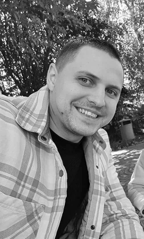 A smiling man with short hair in a plaid shirt and black t-shirt, sitting outdoors with trees in the background.