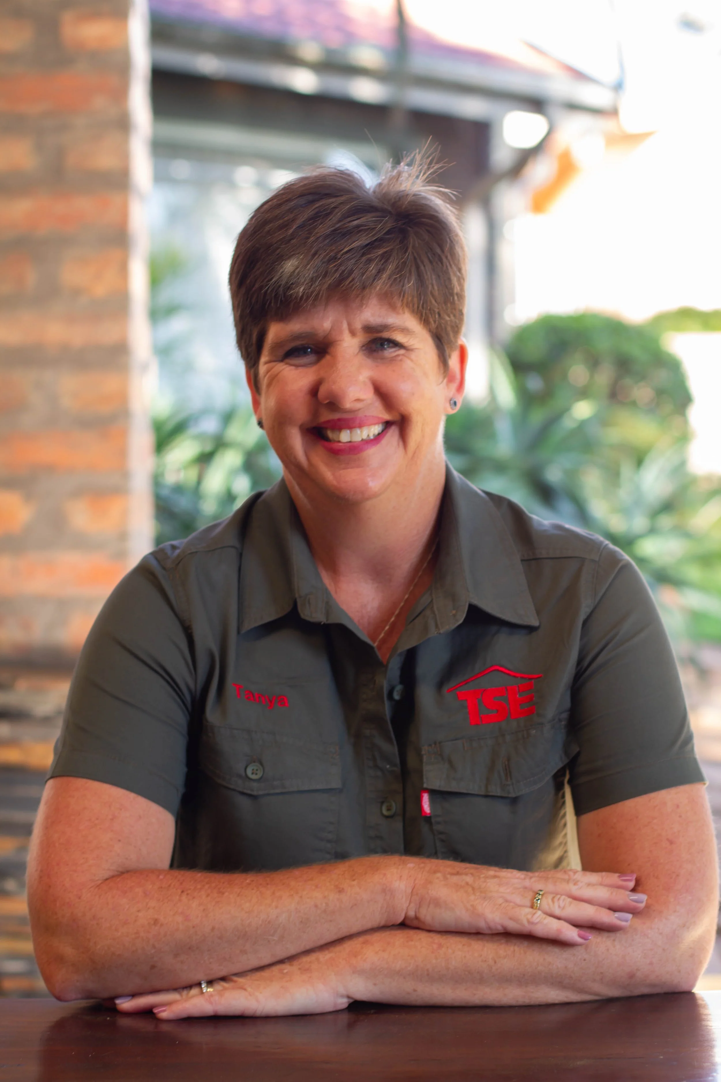 A smiling woman with short brown hair, wearing a gray shirt with red embroidery that says 'Tanya' and 'TSE,' sitting at a table with arms crossed, in a bright setting with a brick wall and green plants in the background.