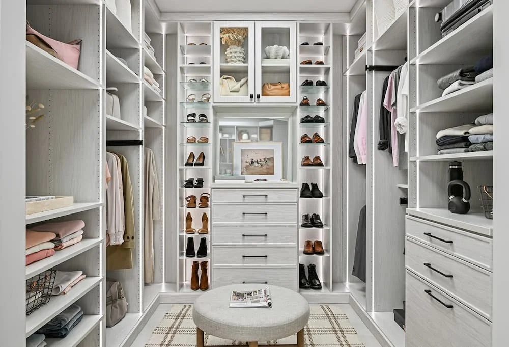 A walk-in closet with white shelves filled with folded clothes, shoes, handbags, and accessories. A small ottoman with a magazine sits in the center, and a rug lies on the floor.