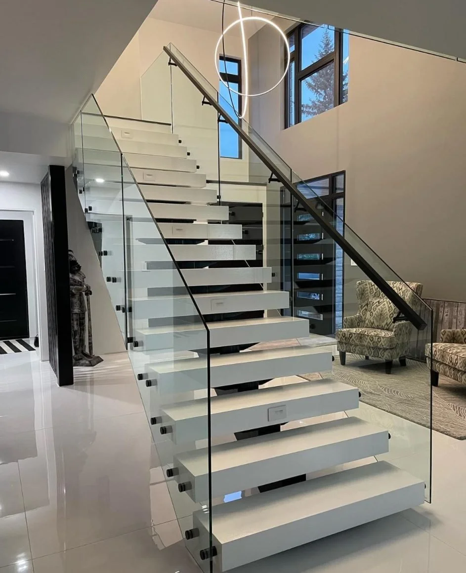 Modern interior staircase with white steps, glass railing, black handrail, and large windows in a well-lit living space.