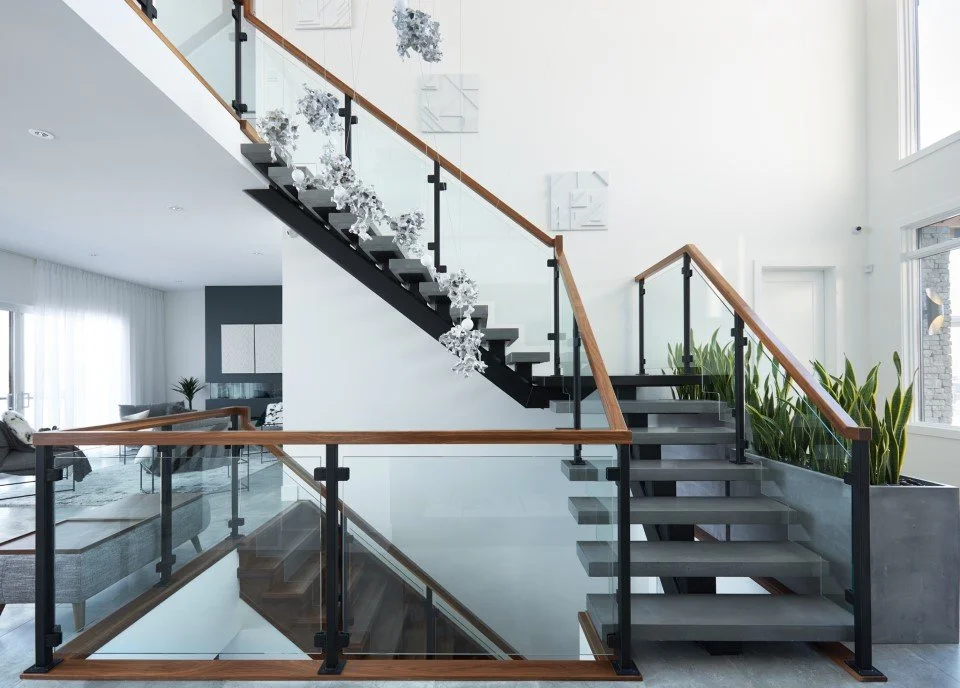 Interior view of a modern stairway with wooden handrails, black metal supports, glass panels, and some large green plants in a spacious, well-lit home.