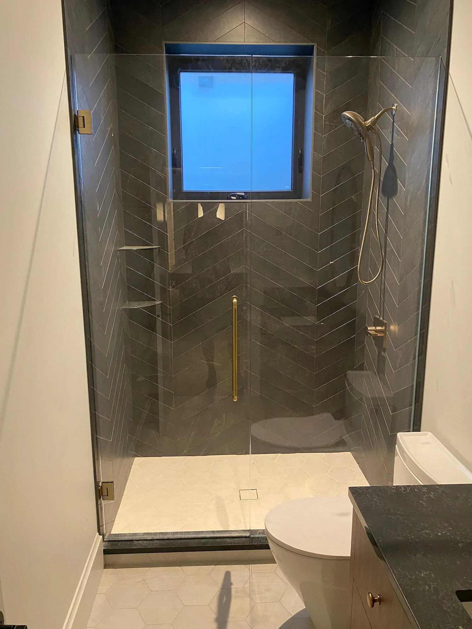 A modern bathroom with a glass shower enclosure, dark tiling, a window, and a toilet.