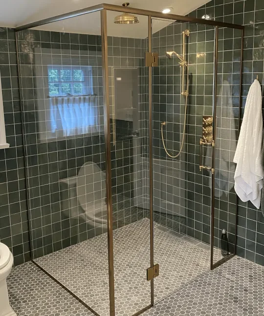 A modern walk-in shower enclosed with glass doors and gold fixtures, located in a bathroom with dark green tiled walls and small hexagonal floor tiles, with a towel hanging on a hook on the wall.