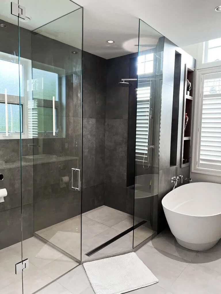 Modern bathroom with glass-enclosed shower, dark tiled walls, a white freestanding bathtub, and large windows with white shutters.
