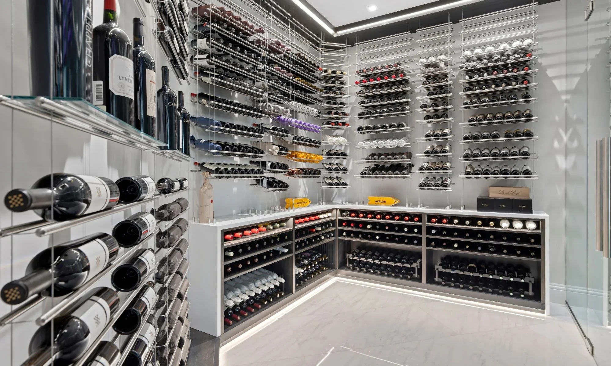 A modern wine cellar with multiple bottles of wine stored on sleek, metal wine racks on white walls and cabinets, with fluorescent lighting.
