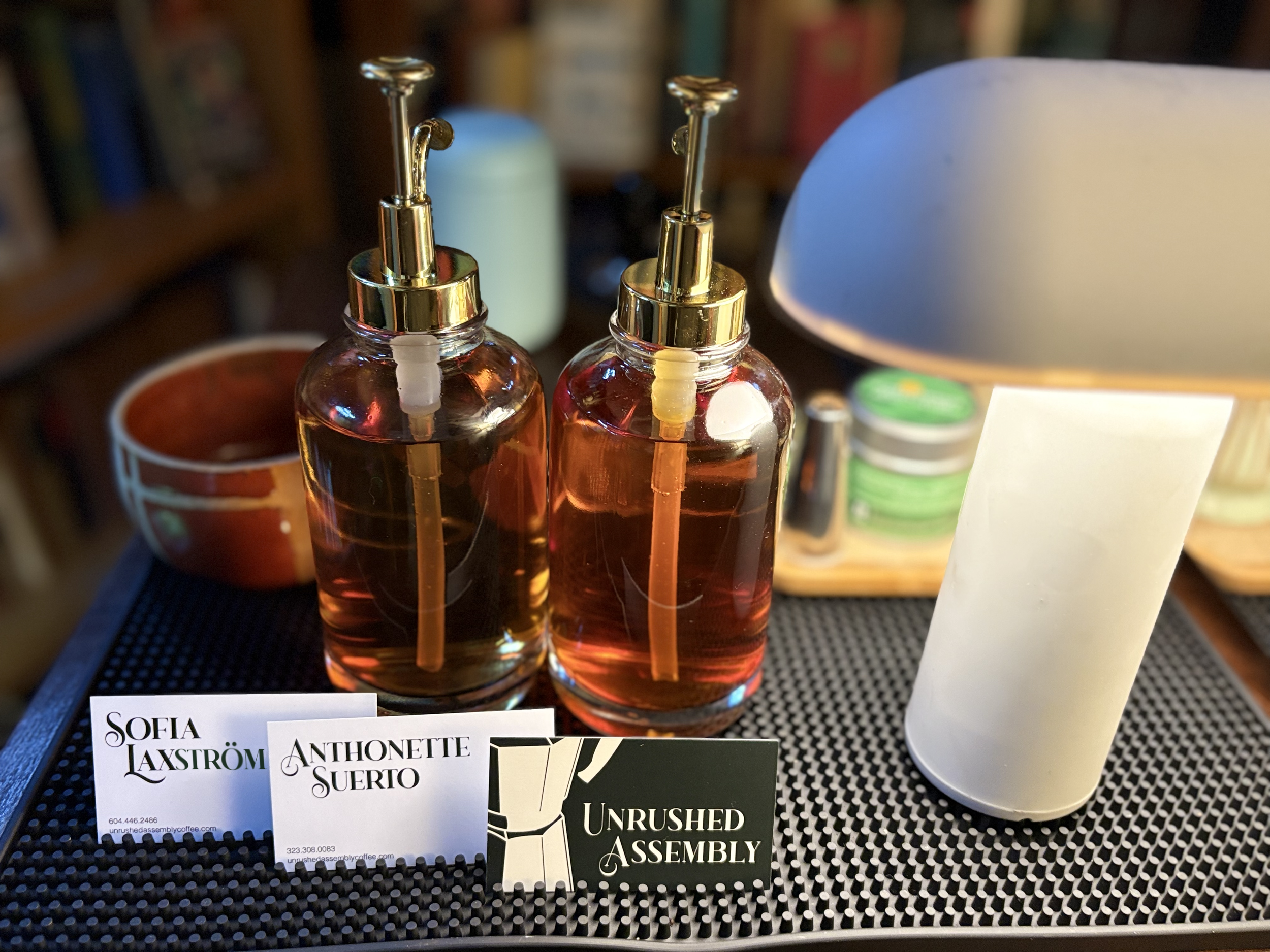 Close-up of two syrup-filled glass bottles with gold-colored spray nozzle caps on a black, bar matt. In the background are their eloquently decorated business cards and a simple lamp, with a blurred bookshelf behind.