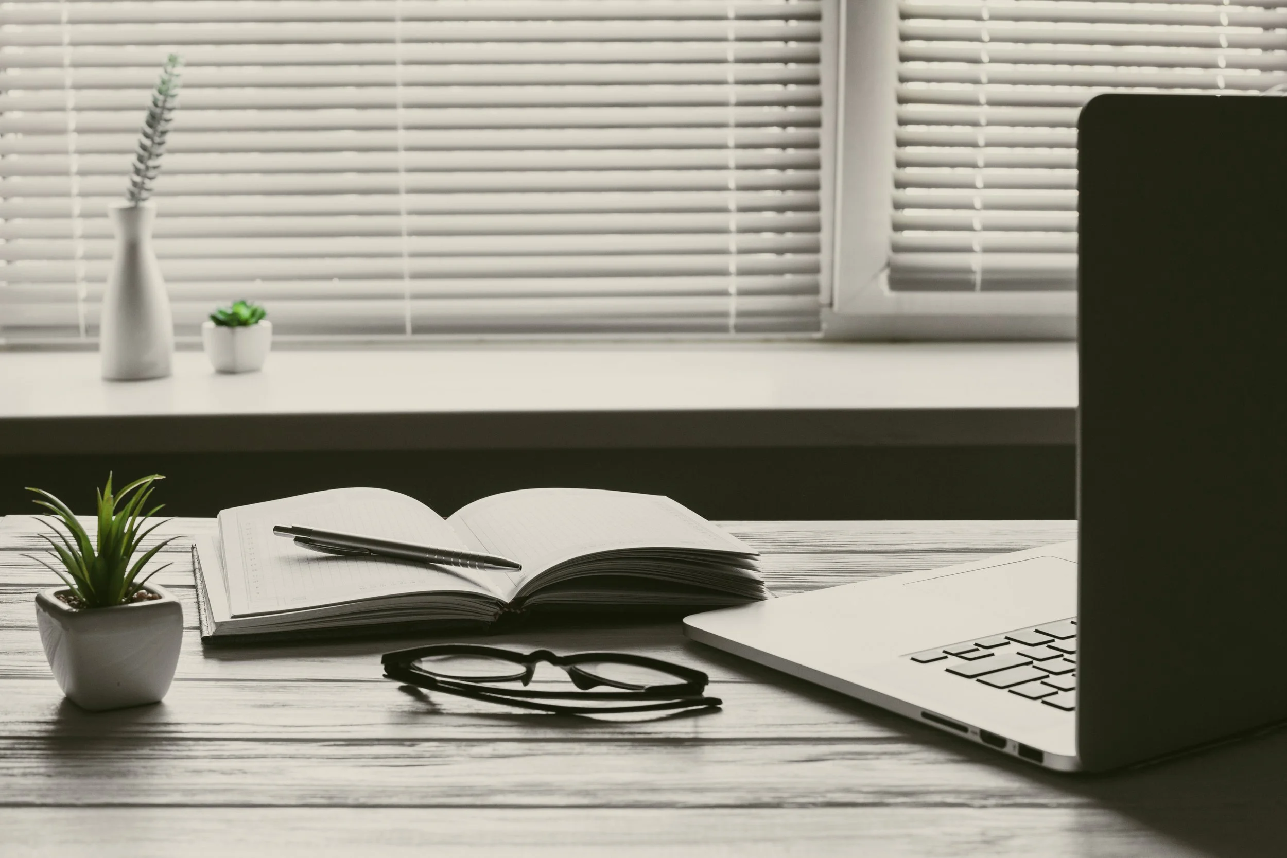 Desk with open notebook, pen, glasses, potted plant, laptop, and decorative vases in front of a window with closed blinds.