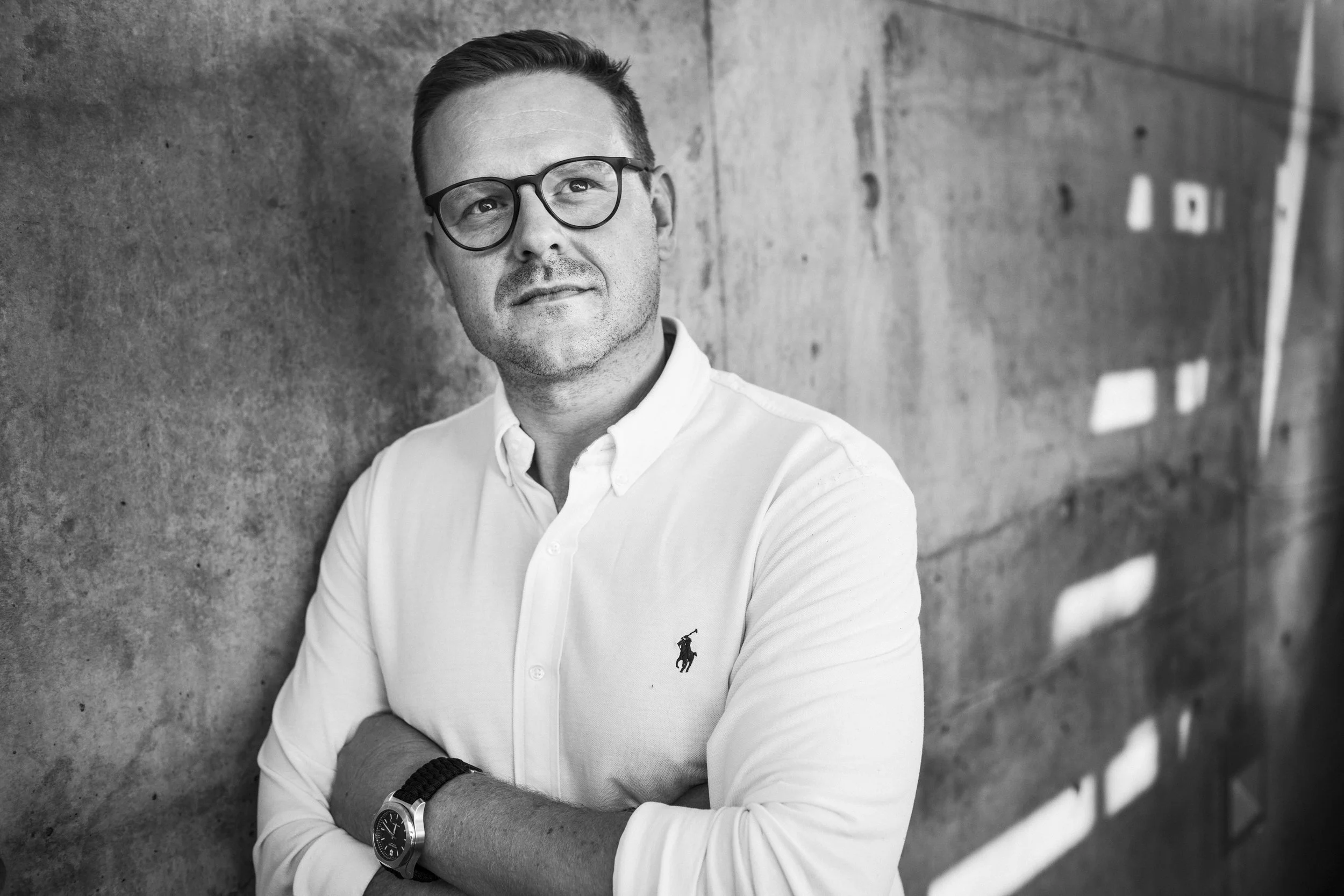 Schwarz-weiß-Fotografie eines Mannes mit Brille, weißem Hemd mit Polo-Logo, Armbanduhr, lässig an eine Betonwand gelehnt.