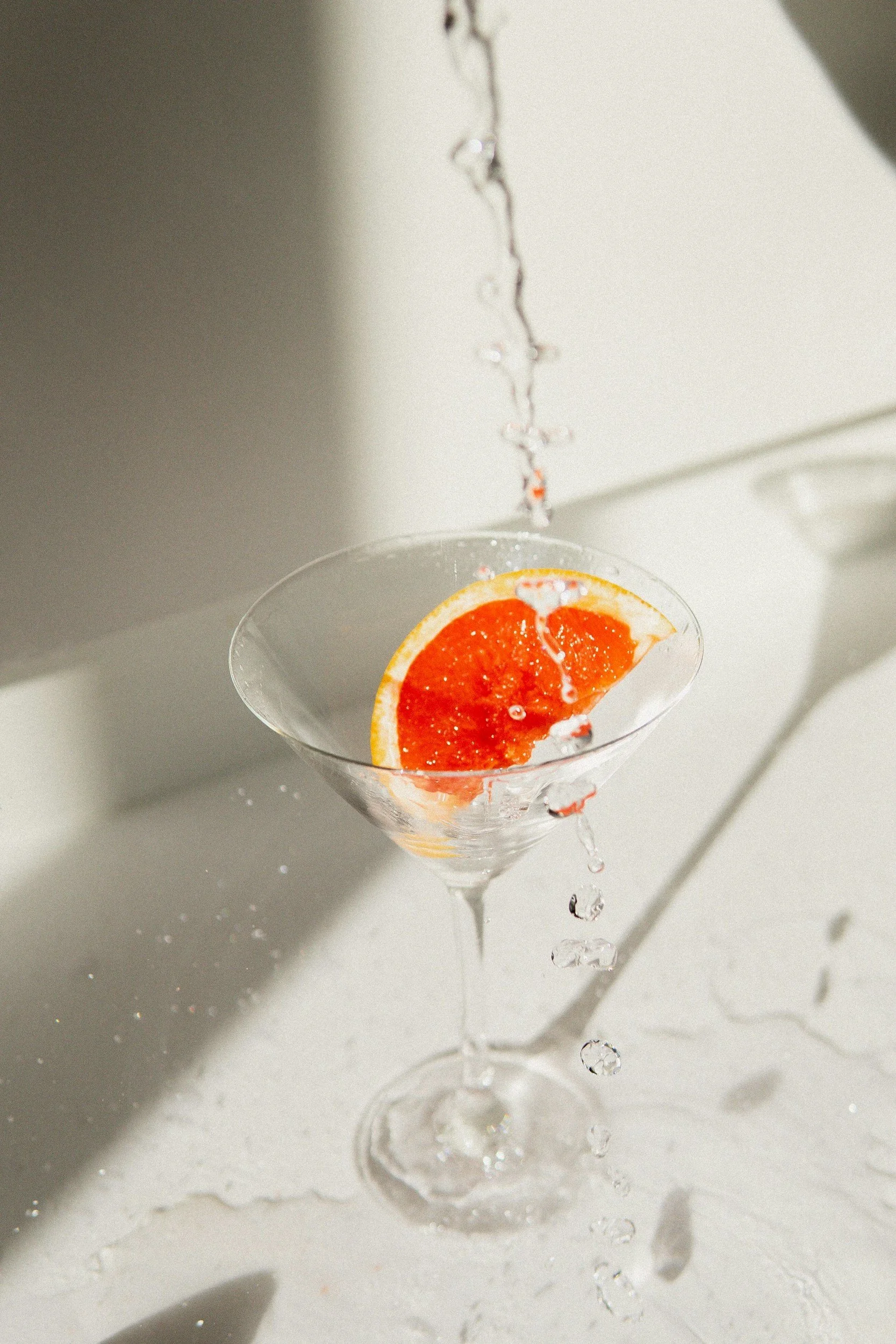 Grapefruit auf einem Martini-Glas, Wasser wird darüber gegossen, mit geteiltem Grapefruit an der Seite, heller Hintergrund.