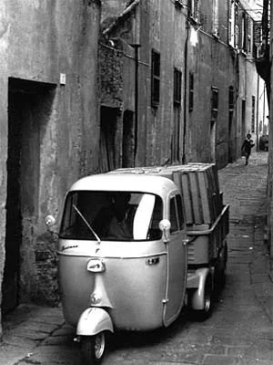 Ein kleiner weißer Motorroller steht auf einer schmalen, alten Straße mit grauen Gebäuden.