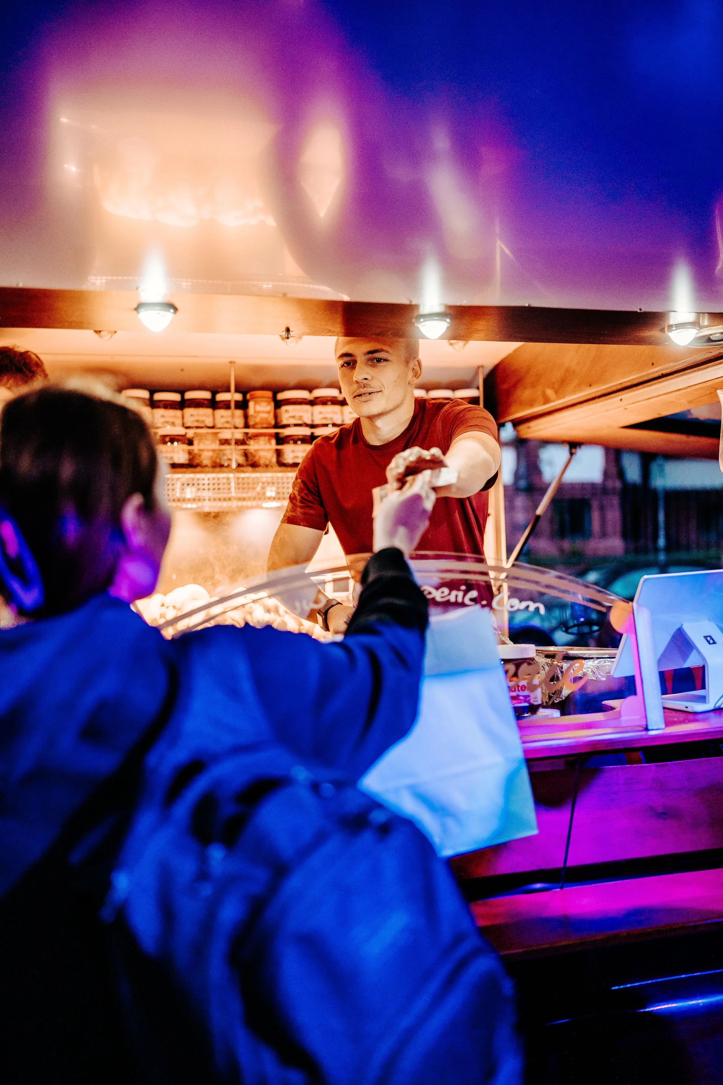 Ein Mann in einem roten T-Shirt verkauft Essen an eine Frau an einem mobilen Imbissstand bei Nacht.