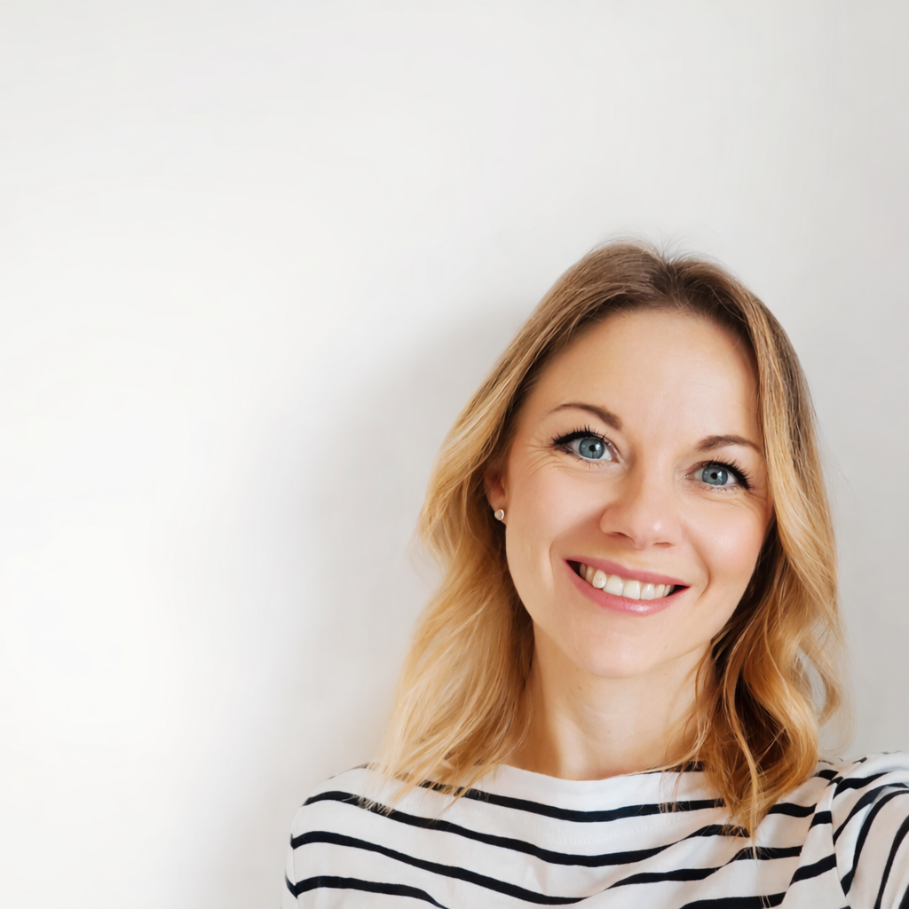A smiling woman with blonde hair, blue eyes, and wearing a white and black striped shirt, standing against a plain light background.