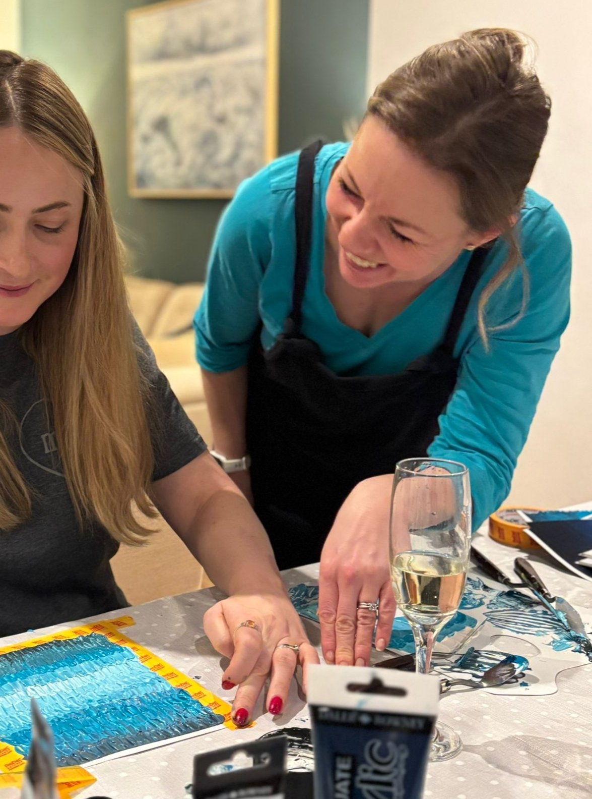 Two women at a table, one pointing at a colorful paint-by-numbers canvas, smiling and the other looking down. There are painting supplies, a glass of white wine, and a marker on the table.