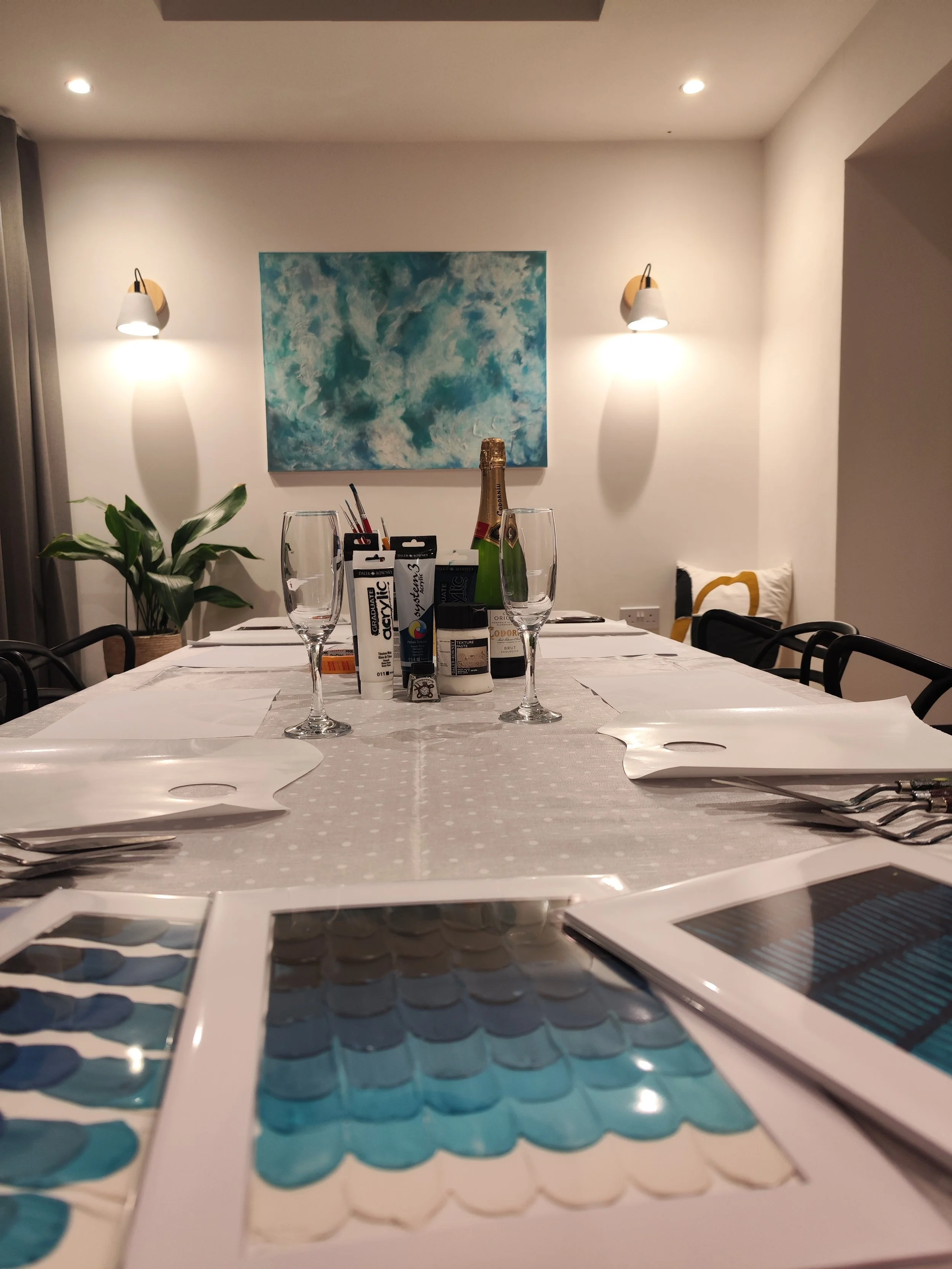A dining table set with white placemats, glassware, and art supplies, in a room with a large abstract blue and white painting and a potted plant on the side.