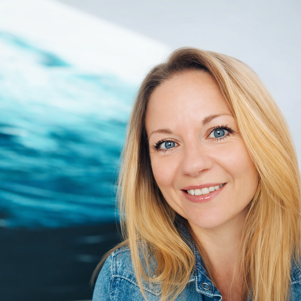 A smiling woman with blue eyes and blonde hair wearing a denim jacket, standing against a blurred ocean wave background.