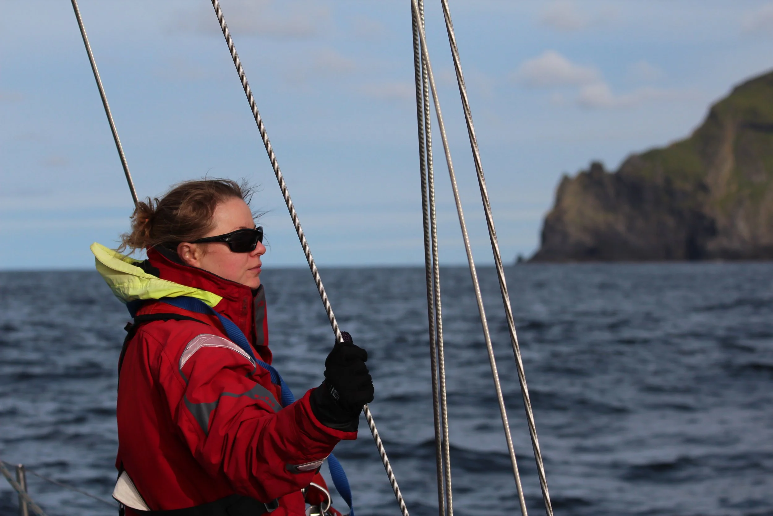 A woman wearing a red waterproof jacket, black gloves, and sunglasses on a sailboat, holding onto the rigging, with a coastal landscape in the background.
