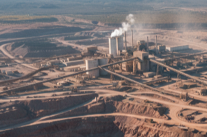 Large industrial or mining facility with smokestacks emitting smoke, surrounded by a desert landscape and roads.