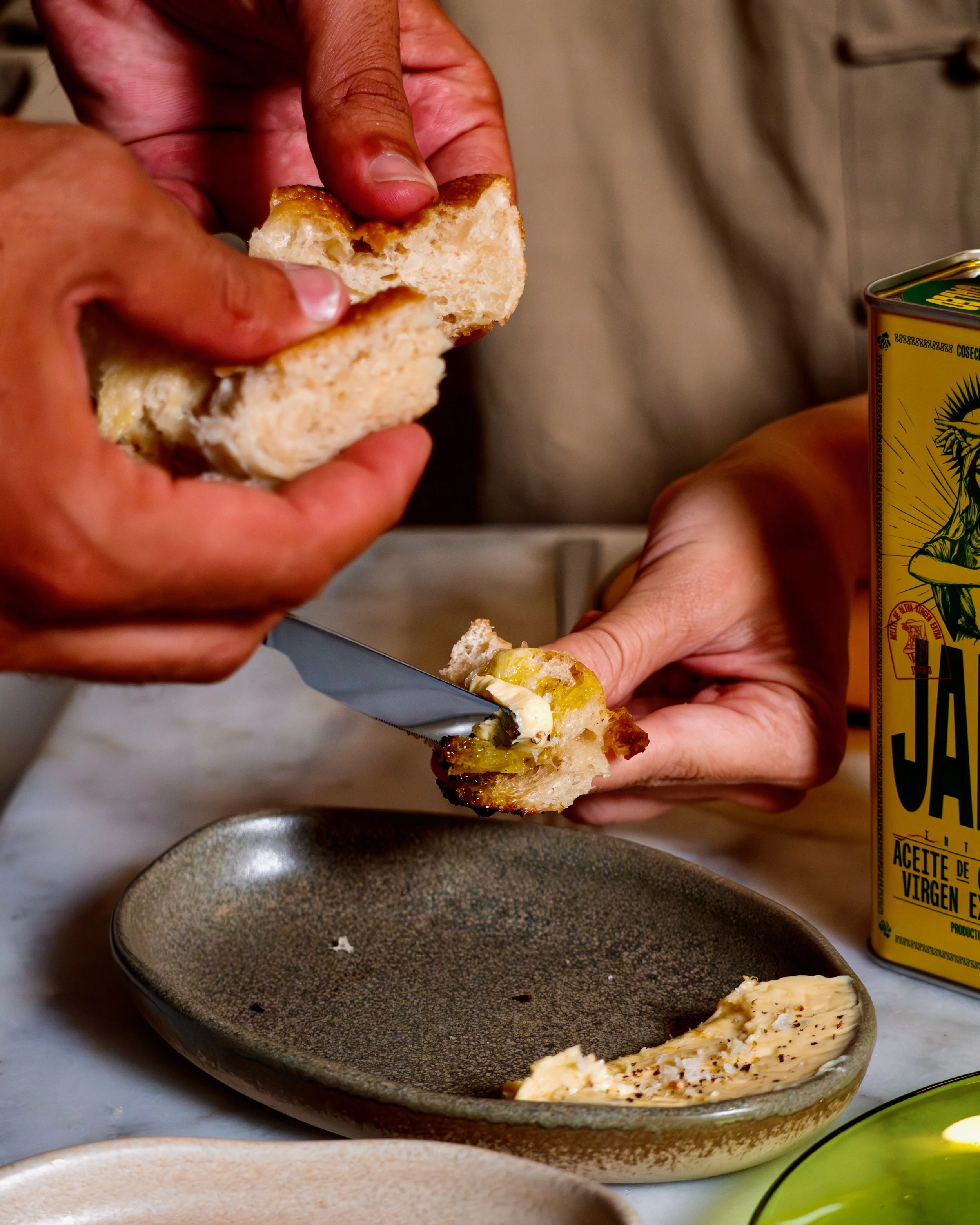 Person breaking apart a piece of focaccia with butter on it over a dish, with a can of olive oil in the background.