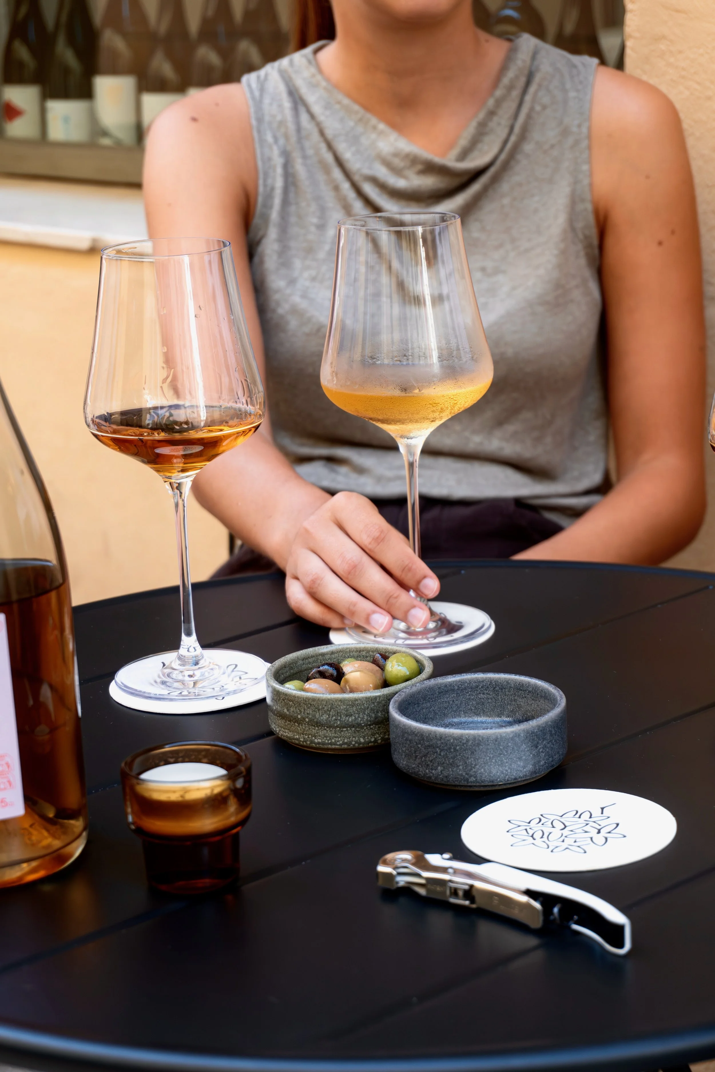A person in a gray sleeveless top sitting at an outdoor table with glasses of wine, small bowls of olives and sauces, and utensils.