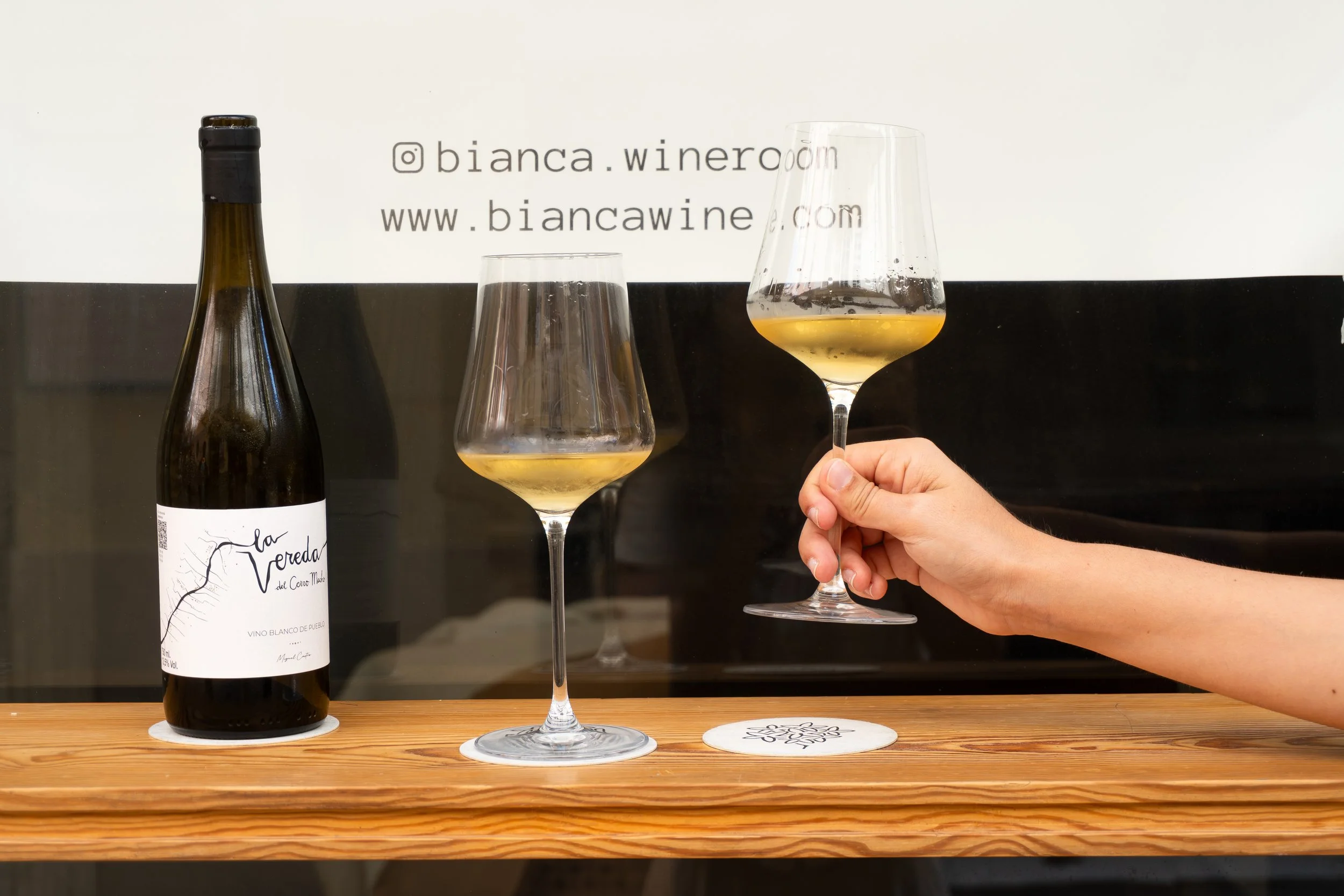 A wine bottle labeled 'La Vereda' and two glasses of white wine, one held by a person's hand, on a wooden table with a white background displaying Instagram and website information for Bianca Wine Room.
