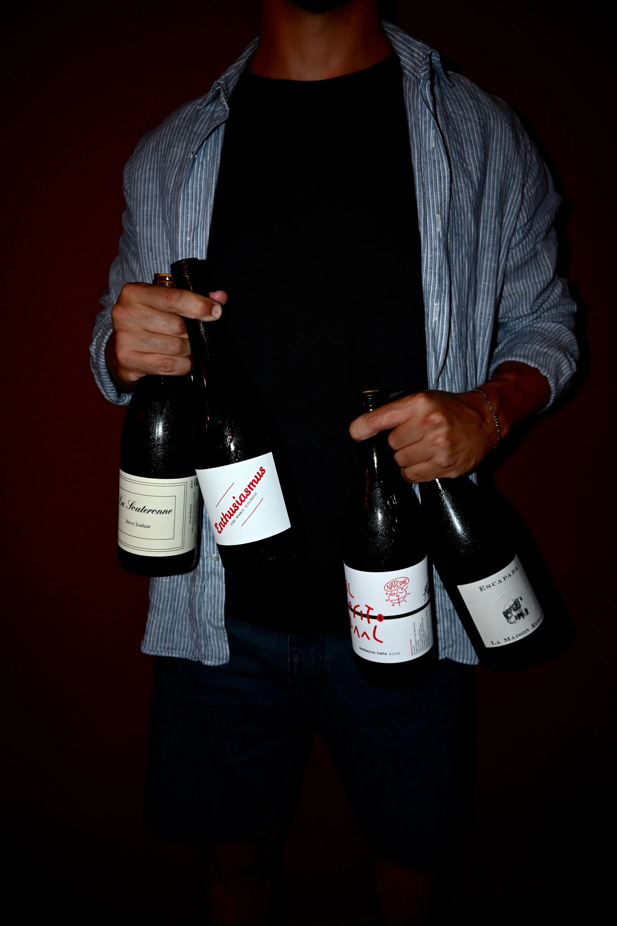 Person holding four wine bottles, standing against a dark background, wearing a black shirt and a blue striped shirt.