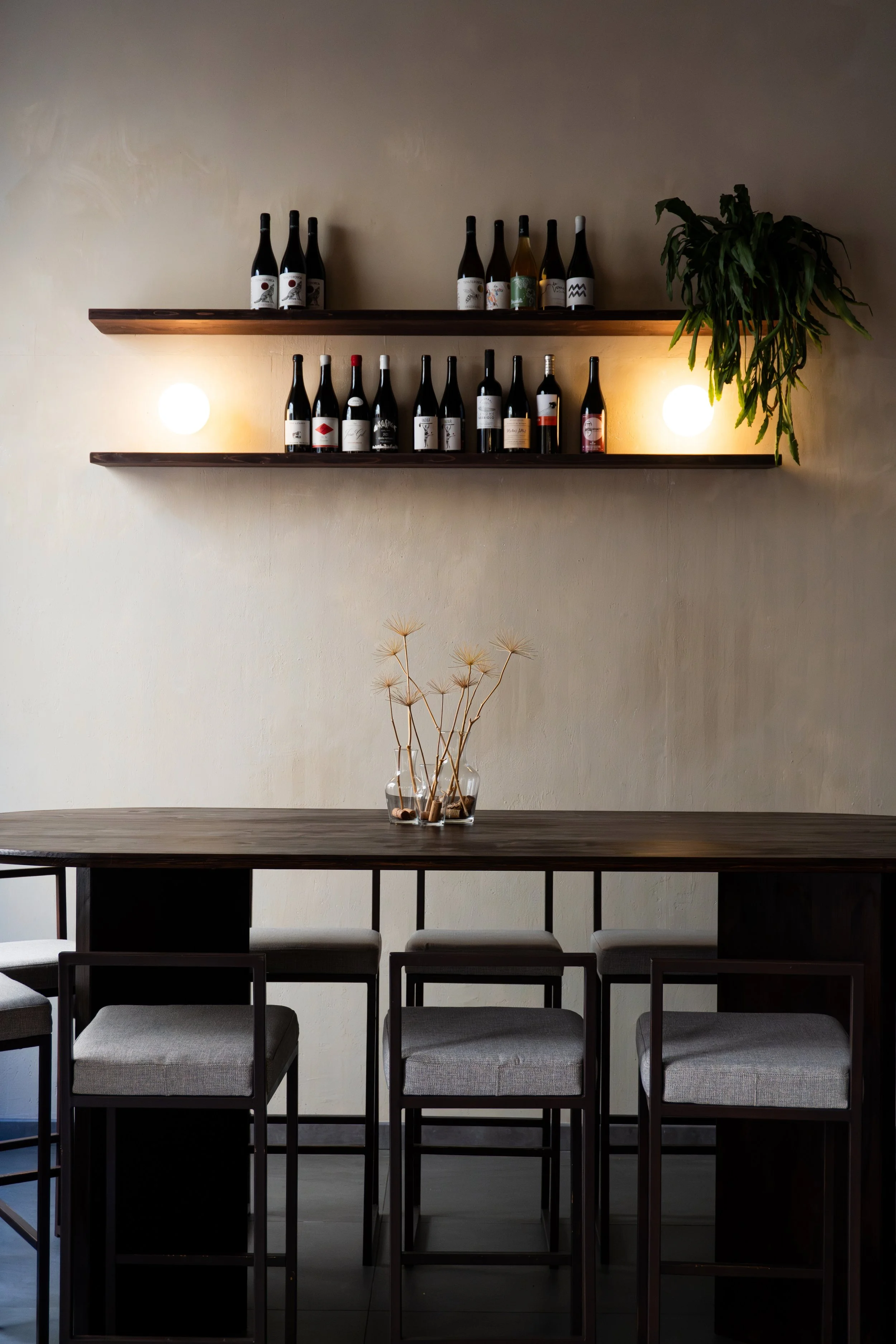 Interior of a dining area with a dark wood table, gray upholstered chairs, and two floating shelves on a beige wall. The shelves hold wine bottles and there are two wall-mounted lights and a potted plant to the right.