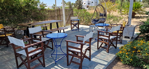 Εξωτερική βεράντα με ξύλινες και μεταλλικές καρέκλες, τραπέζια και μια κρεμαστή καρέκλα σε σχήμα αυγού, περιτριγυρισμένη από φυτά και λουλούδια.