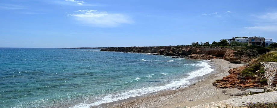 A rocky beach with clear blue water and a partly cloudy sky, with modern houses on a hill in the background.