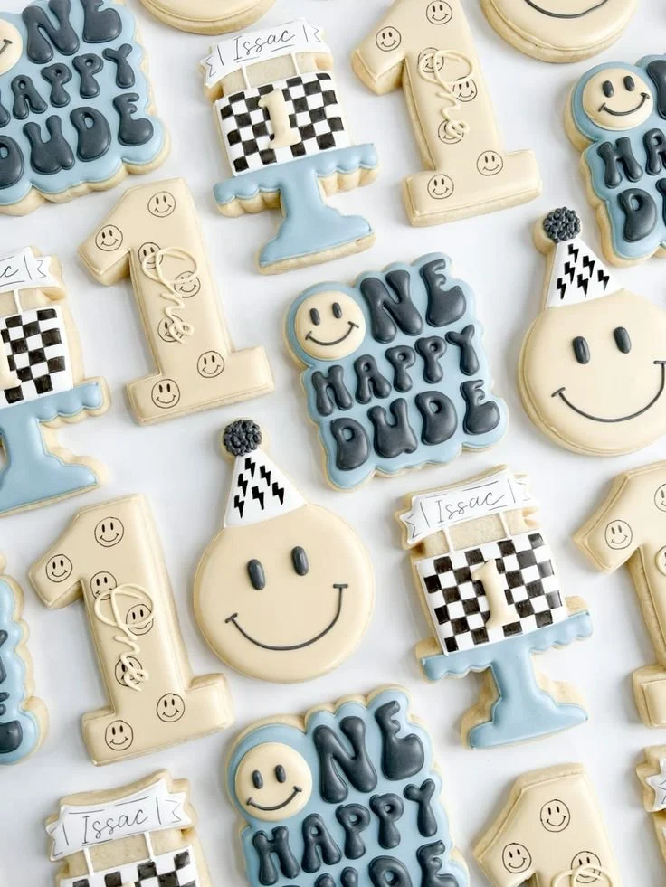 Decorative cookies with birthday and celebration theme, featuring smiley faces, first birthday numbers, and congratulatory messages in blue, beige, and black icing.