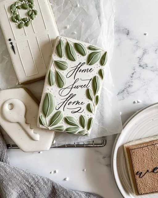 Decorative cake with the words "Home Sweet Home" written on it, surrounded by green leaf-shaped decorations, resting on a marble surface with a white plate and book nearby.