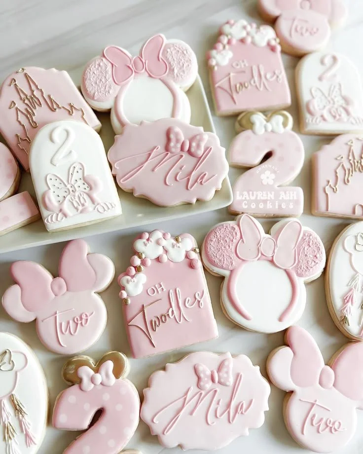 An assortment of pink and white decorated cookies with themes of Minnie Mouse, number two, and 'Oh Toodles' message, arranged on a white tray.